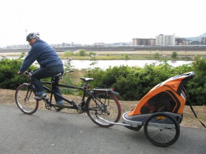 （写真）２人乗りのタンデム自転車にチャイルドトレーラーをつけて、武庫川河川敷をコース点検のため試走しているところ。試走なので、一番前しか人は乗っていいません