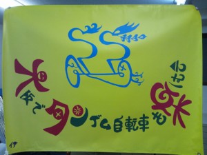 （写真）旗の写真。地色は黄色、タンデム自転車は青色、大阪でタンデム自転車を楽しむ会の文字は緑色で、そのうち、大、タン、楽はショッキングピンクです。