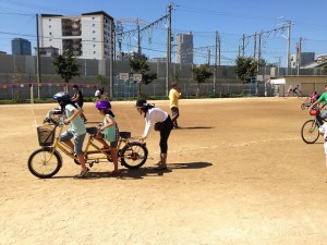 （写真）女の子が２人でタンデム自転車に乗っている後ろから大人が支えています
